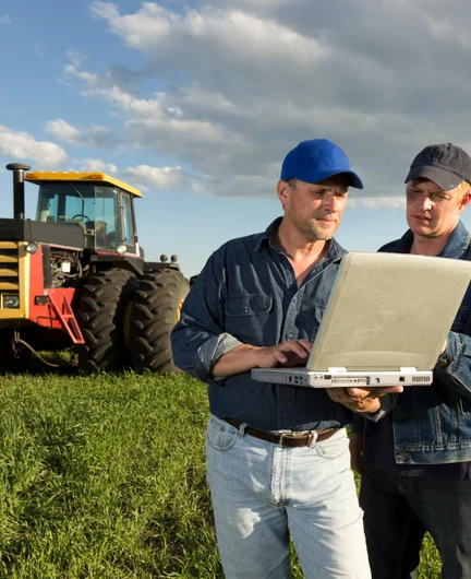 187c0bee-2459-44b0-996c-c71f40947289-two-people-using-laptop-in-the-middle-of-crops-with-heavy-tractor-in-background.jpg
